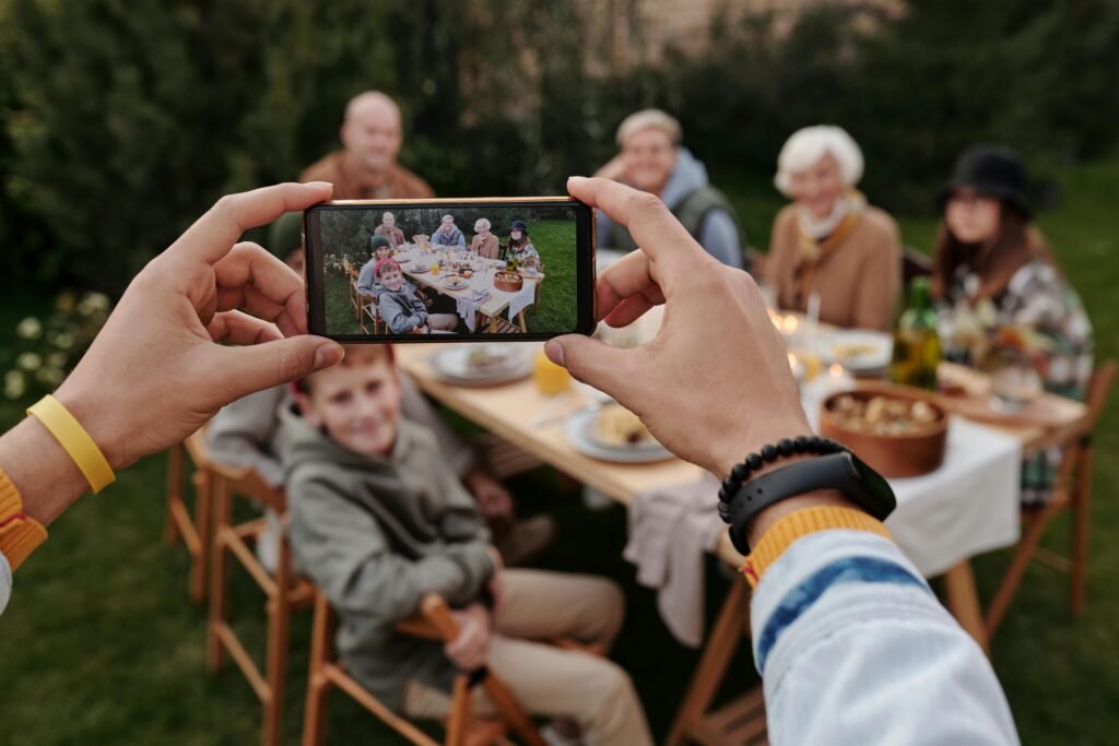 Person taking a group photo of family sitting at an outdoor table using a smartphone.