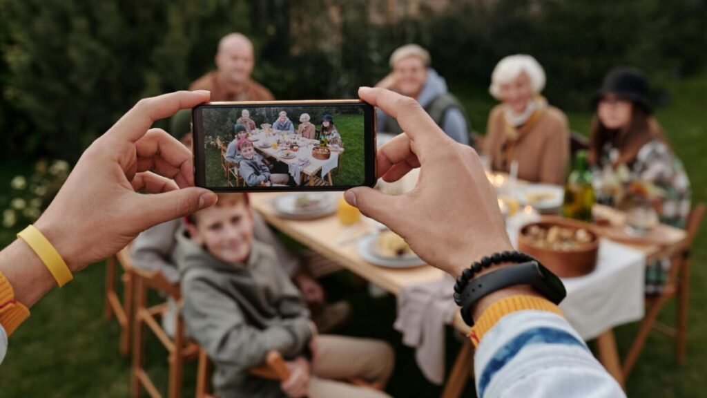 Person taking a group photo of family sitting at an outdoor table using a smartphone.