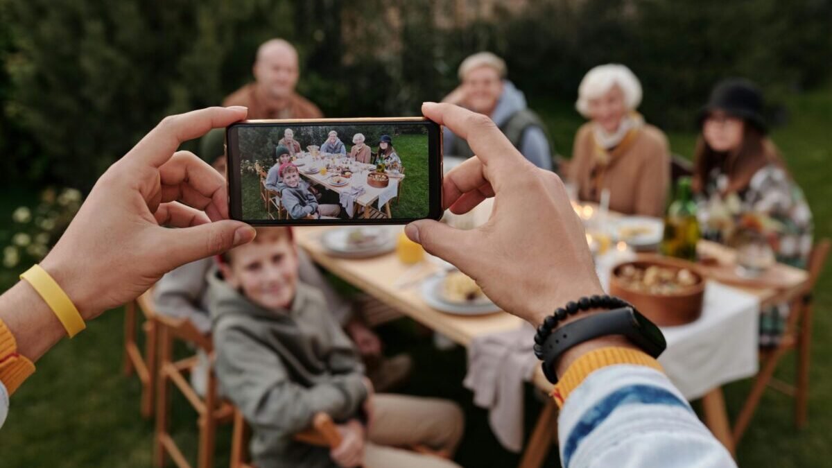 Person taking a group photo of family sitting at an outdoor table using a smartphone.