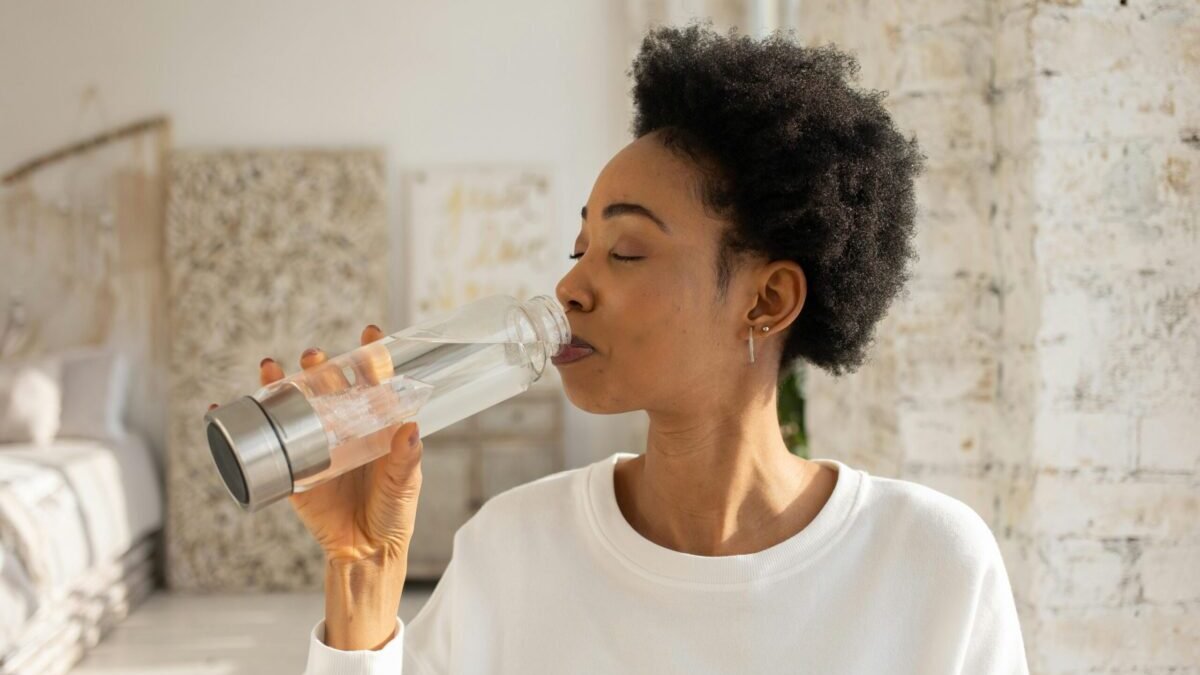 A woman drinking water from a clear bottle indoors, representing healthy hydration habits.