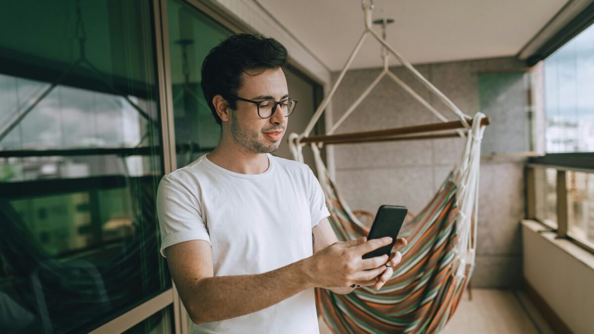 Man wearing glasses using his smartphone while standing on a balcony, with outdoor furniture and a relaxed setting in the background.