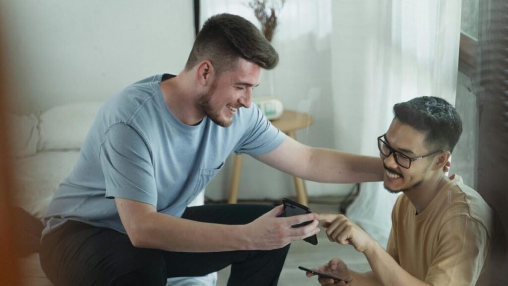 Two men sitting together indoors, smiling and discussing something on a smartphone.